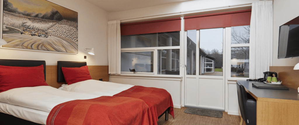 Hotelværelse med to enkeltsenge, hvide dyner og røde sengetæpper, stort vinduesparti med terrassedør samt skrivebord og tv.