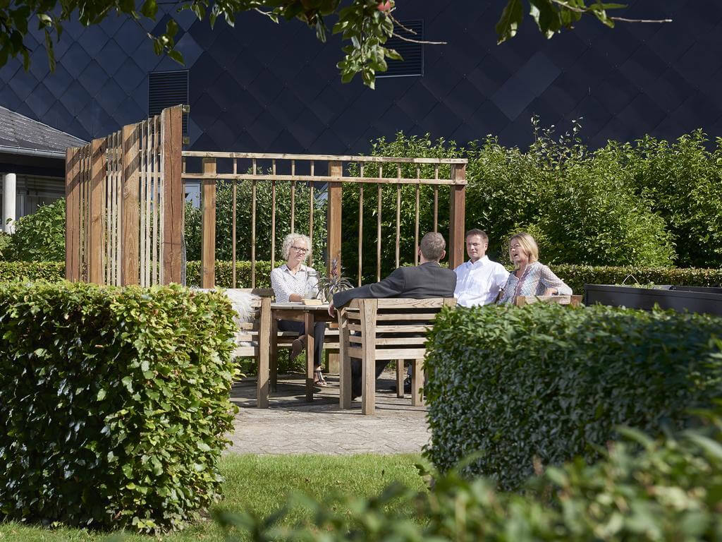 Fire personer sidder og taler ved et havebord p&aring; en terrasse omgivet af gr&oslash;nne buske og tr&aelig;v&aelig;rk i solen.