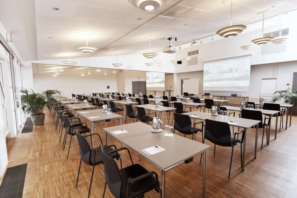 Stort, lyst kursus- og konferencelokale med borde og sorte stole i rækker, vandkander og notatark på bordene samt projektorlærred foran.