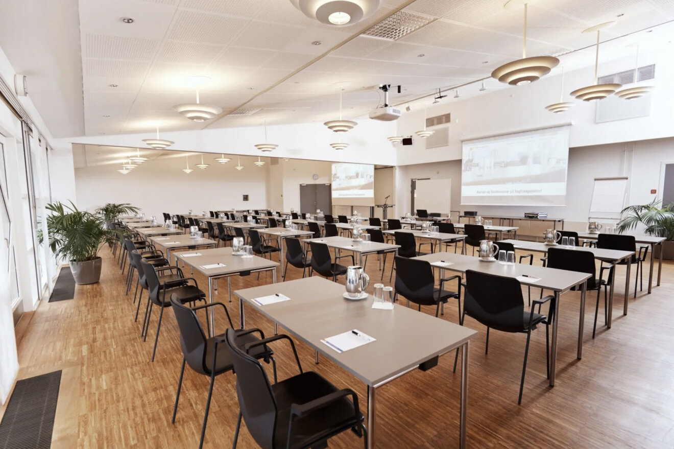 Stort og lyst konferencelokale med borde og sorte stole i rækker, vandkander og notatpapir på bordene samt projektor og lærred til præsentation.