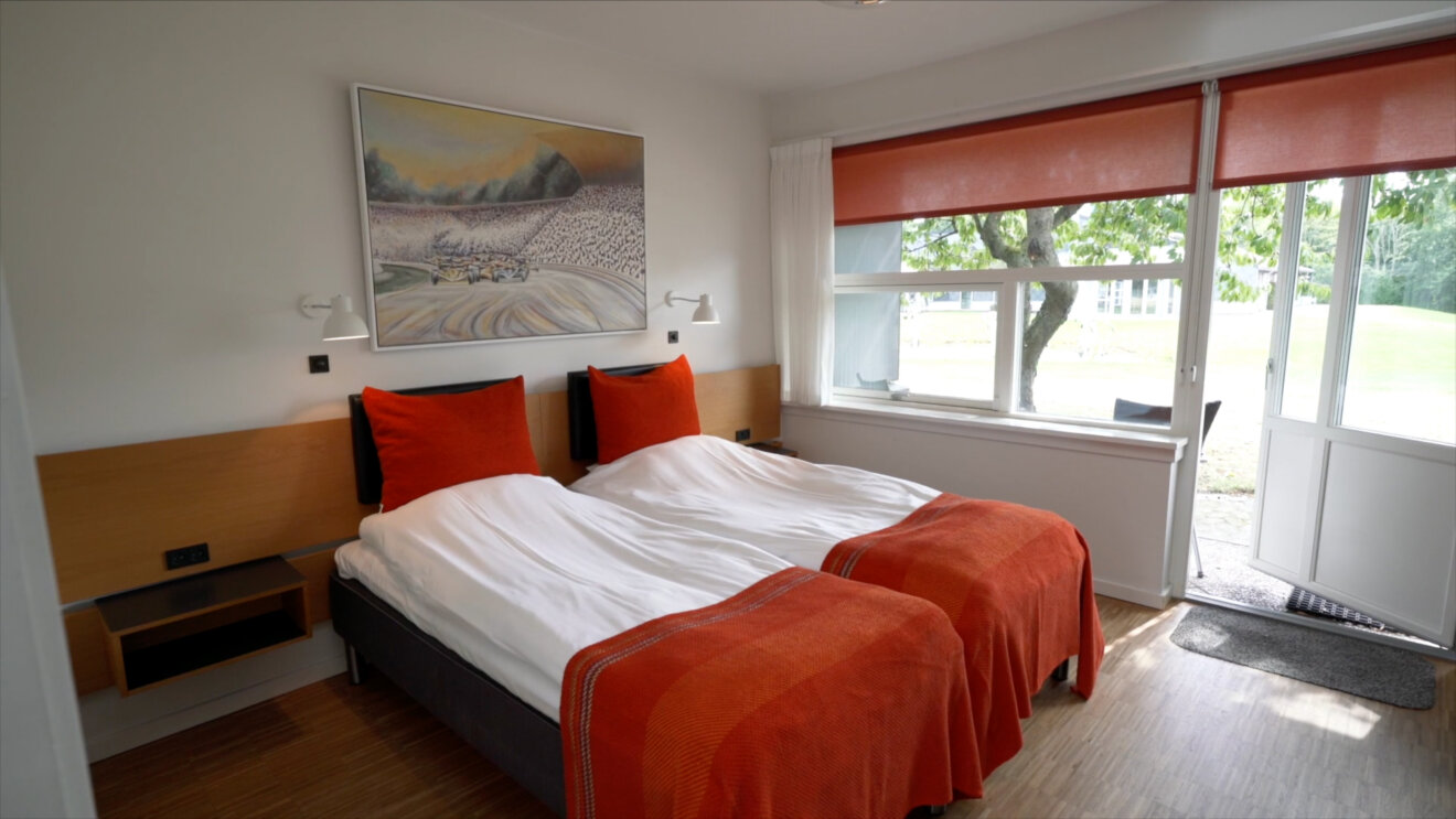 Hotelv&aelig;relse med dobbeltseng, hvide dyner og orange senget&aelig;pper, v&aelig;glamper samt stort vindue og terrassed&oslash;r med udsigt til gr&oslash;nne omgivelser.