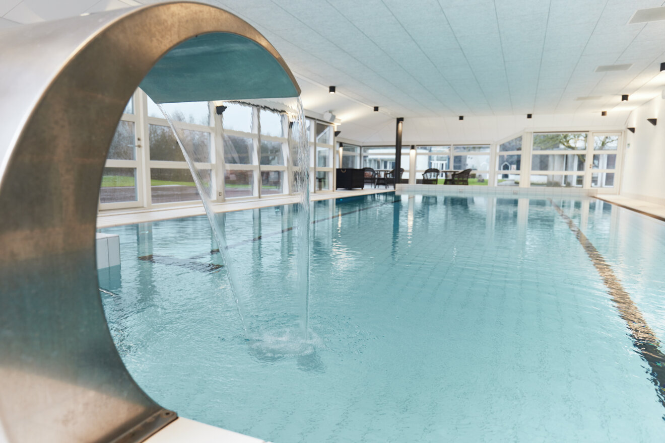 Indend&oslash;rs swimmingpool med en stor svungen vandskulptur i st&aring;l, der sender en bred str&aring;le af vand ned i det lysebl&aring; bassin. Store glaspartier i baggrunden giver kig til gr&oslash;nne arealer.