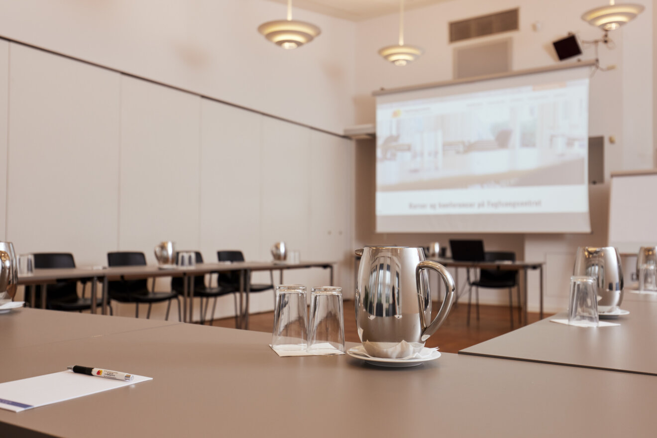 Et lyst og moderne mødelokale klargjort til konference. I forgrunden ses et dækket bord med en vandkande i stål, glas og en blok med kuglepen. I baggrunden ses rækker af borde og stole samt et lærred med en projektion af en præsentation.
