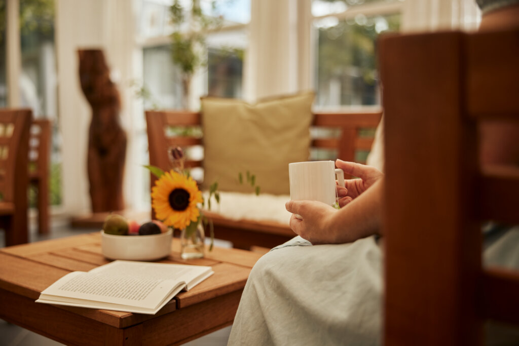 I orangeriet sidder en dame med en kop i hånden, foran hende på bordet ligger en åben bog, en skål med frugt og en vase med en solsikke,