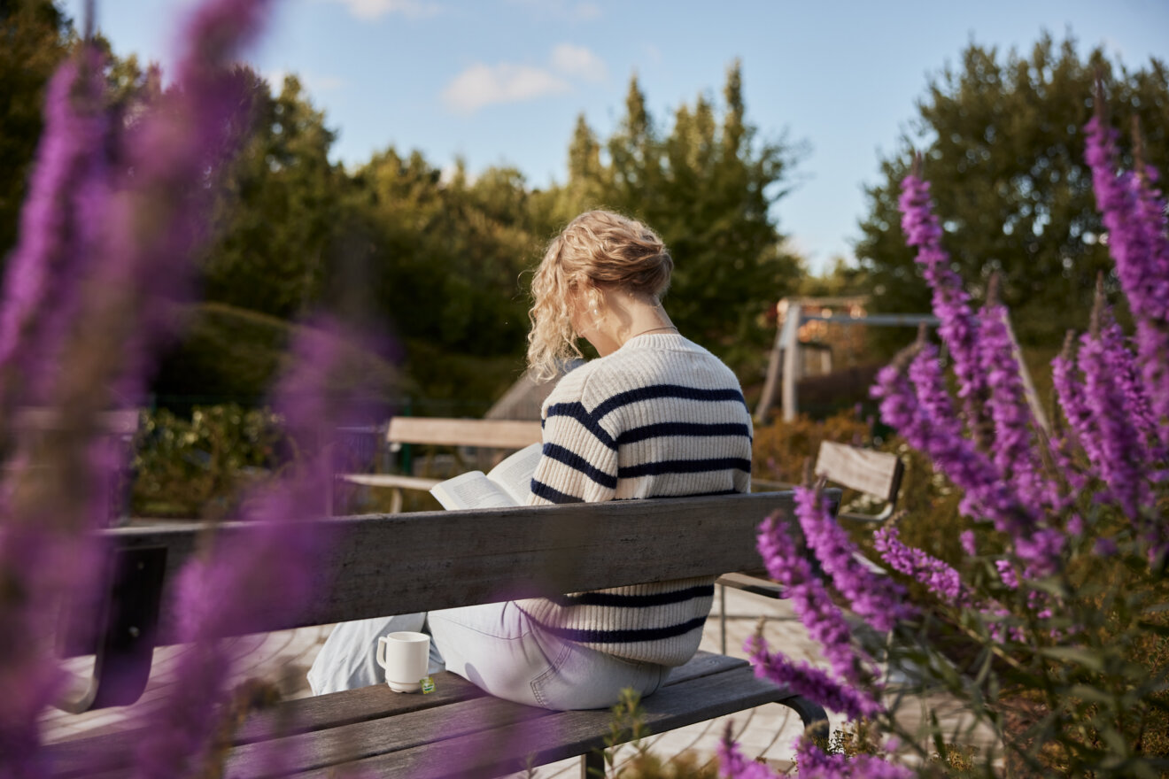 En kvinde sider p&aring; en b&aelig;nk i sansehaven blandt lilla blomster. Hun har en kop te st&aring;ende p&aring; b&aelig;nken ved siden af sig og l&aelig;ser i en bog.