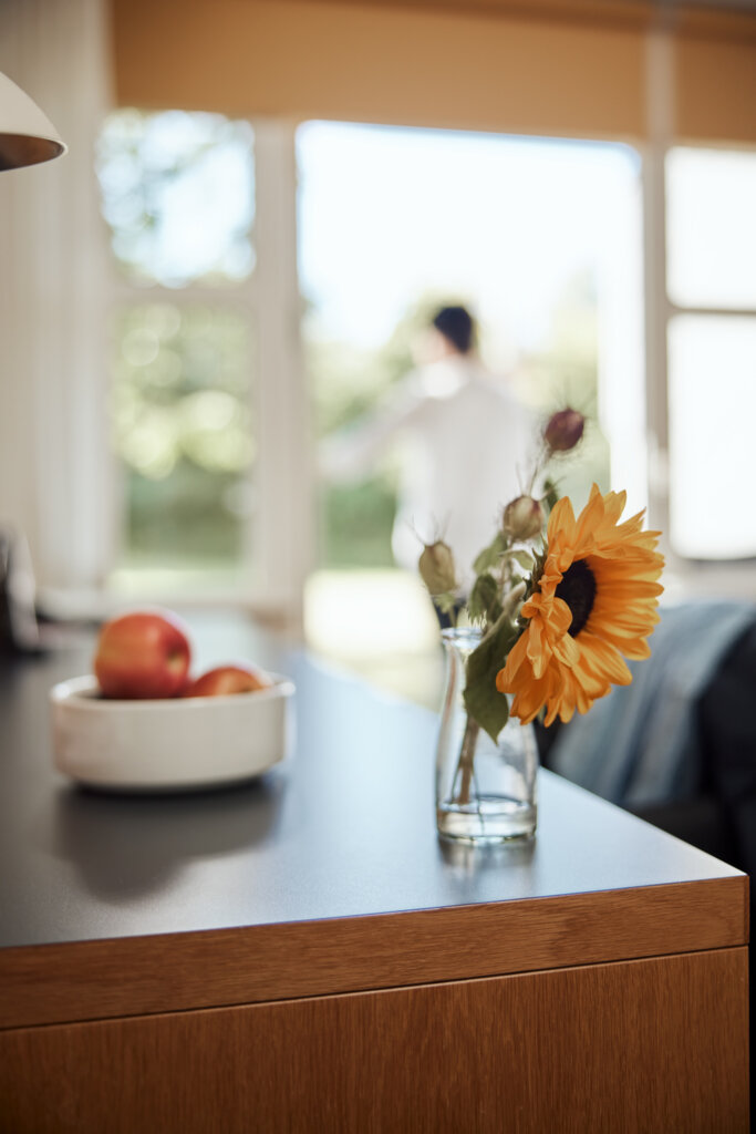 Lille vase med en solsikke står på skrivebordet inde på værelset. ved siden af er der en skål med æbler. I den slørede baggrund anes en person på vej ud af den åbne terrassedør - udgang og kig til grøn natur og dagslys.