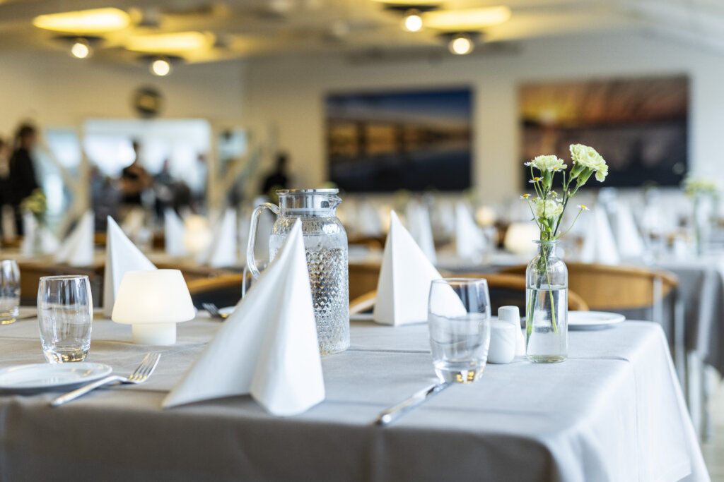 Smuk borddækning med lysegrå duge, hvide servietter og hvidt service. Der er glasvaser på bordene med hvide Nelliker.    
