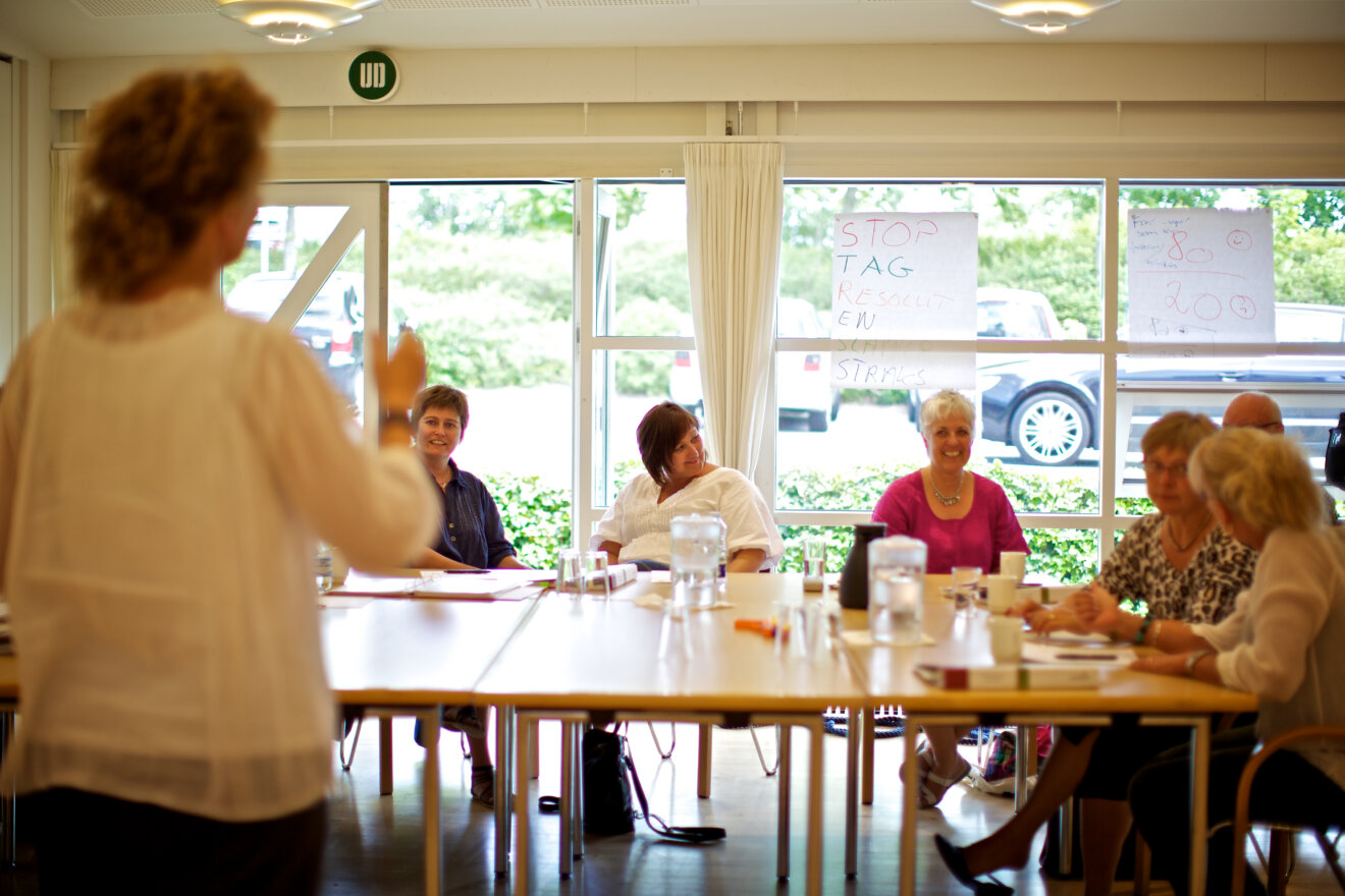 Langs vinduet står en række borde, hvor kursisterne sidder. De kigger på en kvinde, der er ved at facilitere mødet.