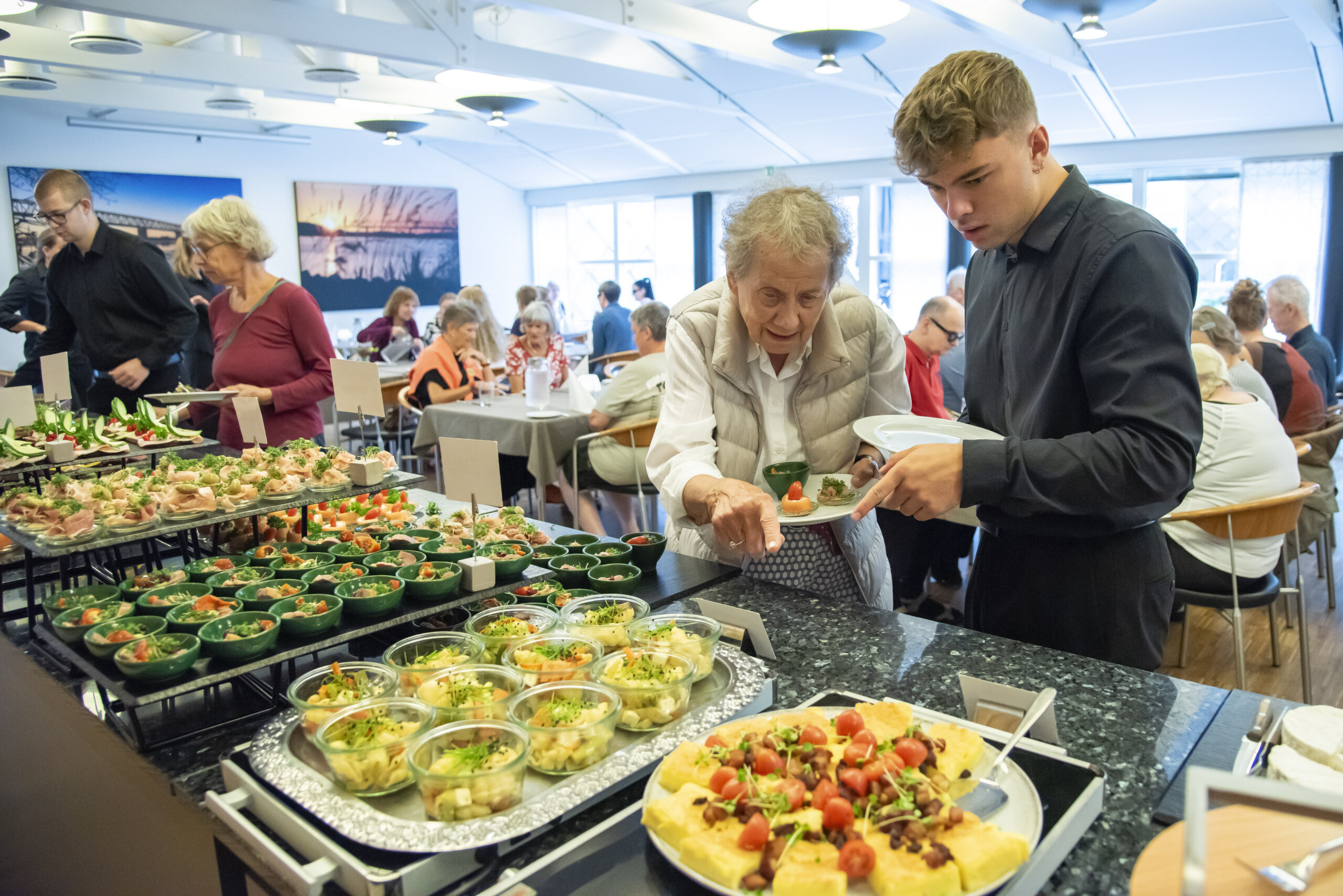 Frokost i restauranten, mandlig tjener står sammen med en ældredame, og hjælper med at tage mad fra buffeten. I baggrunden kan man se andre der spiser og hygger.